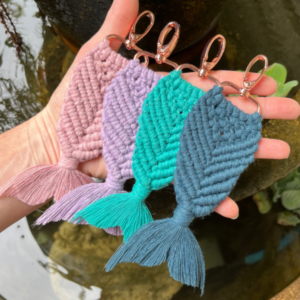 Four macrame mermaid keychains displayed above a fish pond. Colours include vintage rose, lavender, teal, and mallard.